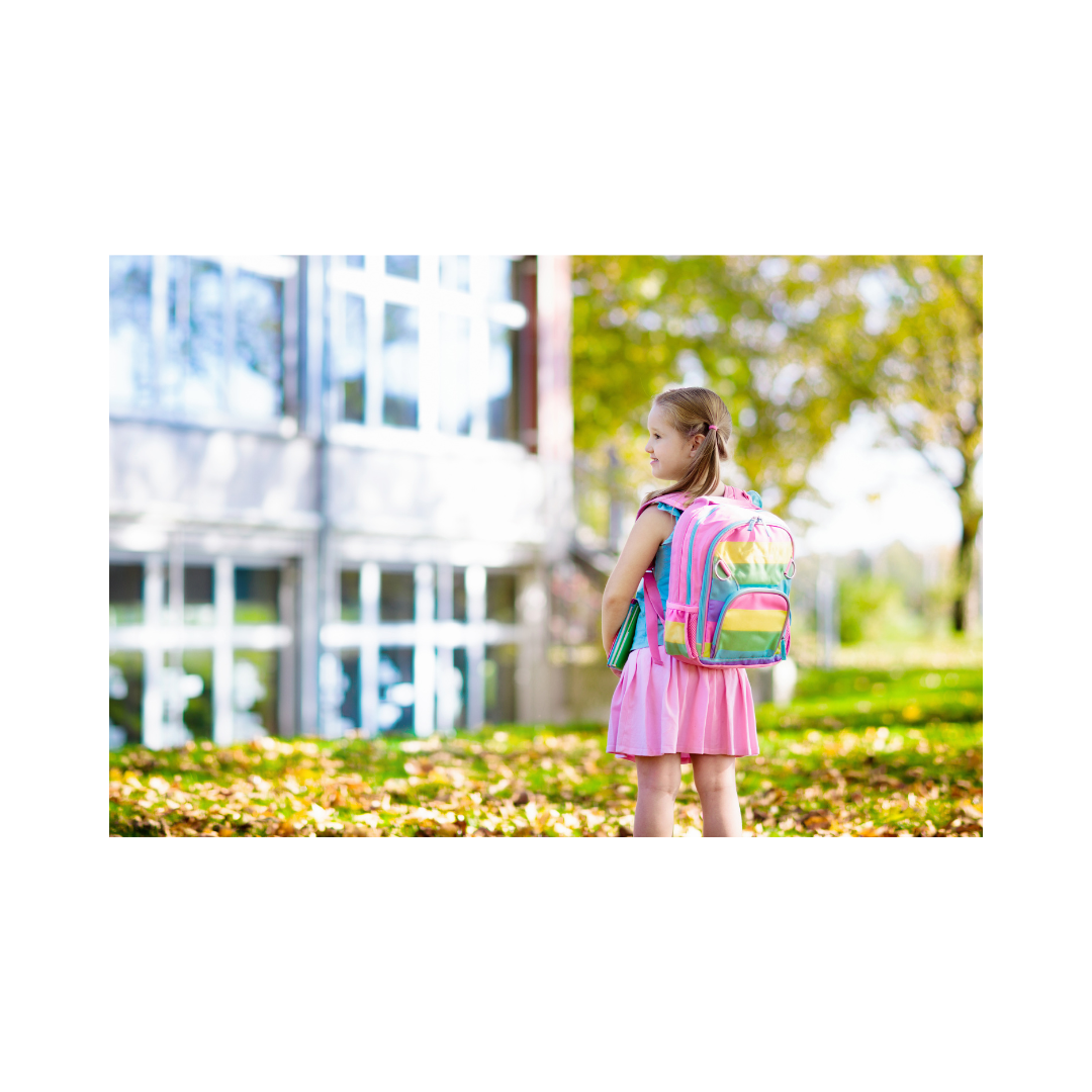 La imagen muestra una niña con su mochila escolar en la espalda, y parada sobre el césped que hay frente a un edificio. Se ve un gran ventanal que entendemos que es la escuela.