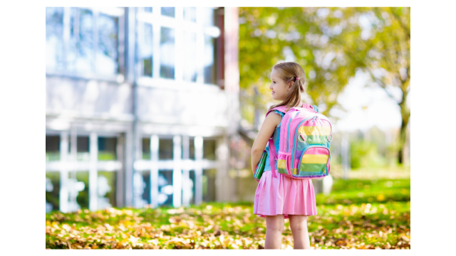 La imagen muestra una niña con su mochila escolar en la espalda, y parada sobre el césped que hay frente a un edificio. Se ve un gran ventanal que entendemos que es la escuela.